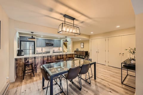 Modern kitchen and dining area with a glass-top dining table, four chairs, a two-tone kitchen with pendant lighting, a wooden island, and light hardwood flooring.