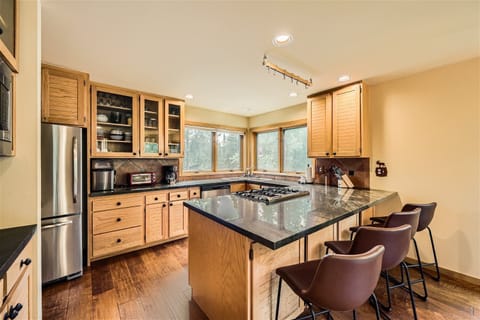 A modern kitchen with wooden cabinets, stainless steel appliances, a black countertop island with a built-in stove, and three brown bar stools against the island. Large windows provide natural light.