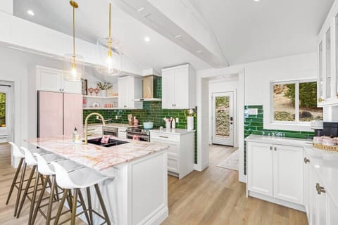 This vibrant kitchen features a pink refrigerator, green tile backsplash, and a marble island with white stools. White cabinets offer ample storage, while gold pendant lights and natural light brighten the space.