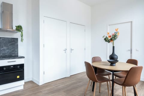 Minimalist dining area with a round wooden table, sleek brown chairs, and a modern black vase centerpiece, framed by clean white walls and doors.