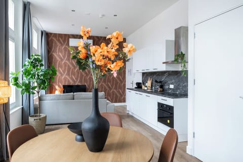 Contemporary dining setup featuring a light wood table, leather-style chairs, and a floral arrangement in a black vase, complemented by a bright, airy backdrop.