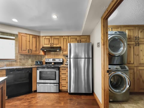 A kitchen with wooden cabinets, a stainless steel refrigerator, an oven, a dishwasher, and a small laundry area with a stacked washer and dryer.