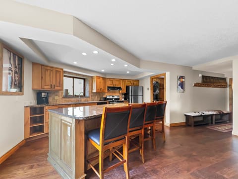 A modern kitchen features a large island with four chairs, wooden cabinets, stainless steel appliances, recessed lighting, and hardwood floors. A bench and coat rack are visible in the background.