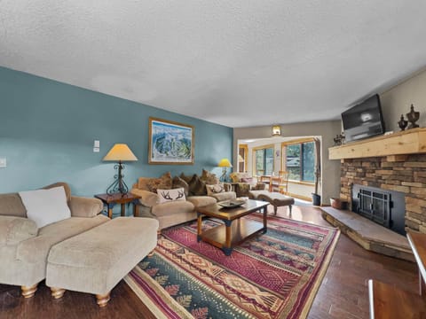 A cozy living room with a couch, armchair, and coffee table on a patterned rug. A stone fireplace sits to the right, and there is a large window and a door leading outside. A painting hangs on the wall.