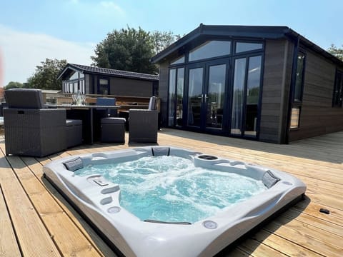 The lodge has entirely glass patio doors that open onto a spacious wooden deck. The deck overlooks a serene outdoor area featuring a sunken hot tub and a four-person dining table. 