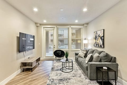 Bright living room w/ cozy sofa seating, wall-mounted TV & large windows letting in natural light