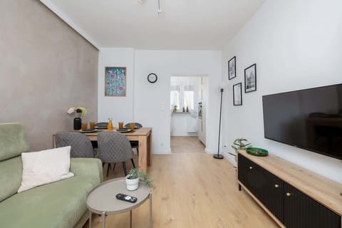 A cozy living room with a green sofa, TV, and dining table, perfect for relaxing or working. Natural light flows through the large windows, creating a bright space.