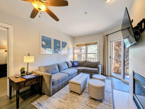 Your inviting living space features a plush sectional sofa, cozy seating, and warm lighting—perfect for unwinding after mountain adventures. Natural light streams through balcony doors, while the ceiling fan keeps you comfortable year-round.