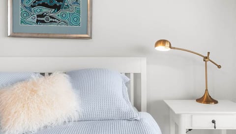 A neatly made bed with a fluffy white pillow and light blue bedding is accompanied by a stylish brass lamp on a white bedside table, with an aboriginal art piece hanging on the wall above the bed.