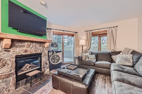 A cozy living room with a stone fireplace, mounted TV, corner sofa, small table, ottoman, window with curtains, and a floor lamp. The room has large windows allowing natural light in.