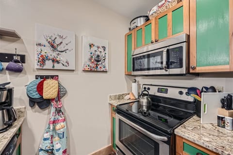 A small kitchen with green and wood-tone cabinets, a stainless steel microwave and oven, granite countertops, colorful potholders, and two animal-themed paintings on the wall.