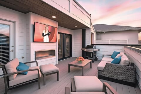 Rooftop patio at dusk with outdoor lounge chairs, gas fireplace, wall-mounted TV, and grill under wooden ceiling
