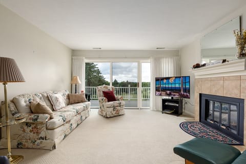 Bright living room w/ fireplace & TV, opening to a balcony overlooking the green golf course.