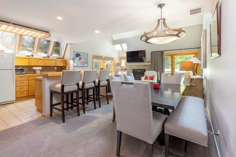 A modern dining and kitchen area with a wooden dining table, upholstered chairs, a kitchen island with bar stools, stainless steel appliances, a fireplace, and a wall-mounted TV.