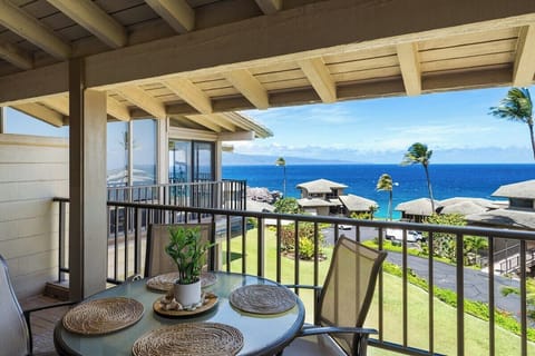 Covered oceanview lanai featuring a round bistro table and chairs, ideal for outdoor dining while enjoying vibrant blue waters, tropical landscaping, and gentle island breezes.
