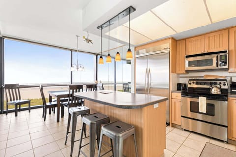Oceanfront cooking and dining with a view! This fully equipped kitchen features an island with bar stools, a dining table, and expansive windows showcasing stunning beach scenery.