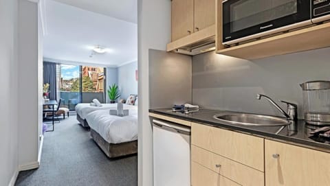A perspective showing a compact kitchen corner with light wood cabinets, an integrated microwave, and a countertop leading into the main bedroom space.