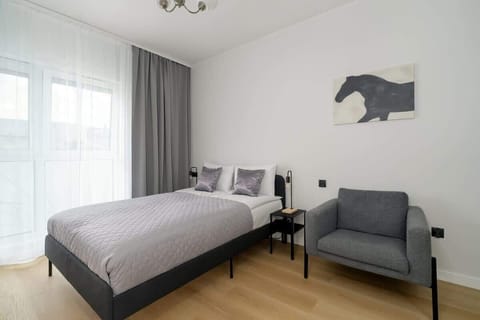 A view of the main area of the studio apartment, featuring a bed, a gray armchair, and large windows with sheer curtains.