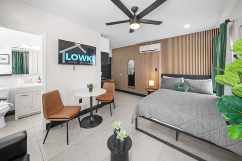 This inviting studio features a cozy queen-size bed, a comfortable leather sofa bed, and stylish decor with vibrant wall art and greenery. Natural light from large windows and a wood-accented wall create a warm, relaxing atmosphere.
