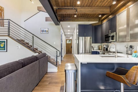 Modern kitchen and living area with wooden ceiling, dark cabinets, stainless steel appliances, and a staircase with metal railing. Cozy seating area with a gray sofa and bar stool.