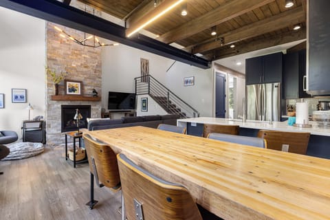 Open-concept living and dining area with wooden ceiling, stone fireplace, large wooden table, and modern kitchen. Stairs lead to another level.