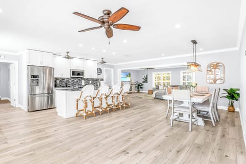 The dining area comfortably seats six beneath a farmhouse chandelier, with four additional rattan counter stools at the kitchen counter. The open floor plan allows for easy serving from the ki