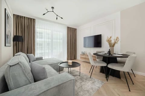 A cozy living room with a gray sofa, coffee table, and dining area in the background. Large windows allow natural light to brighten the space.