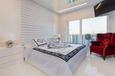 A modern bedroom with a large bed, white interiors, and a red armchair. Floor-to-ceiling windows brighten the room with natural light.
