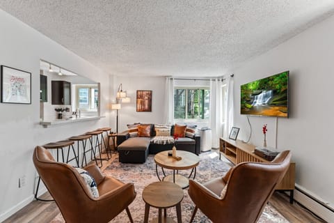 Charming and contemporary living space featuring a sectional sofa, leather chairs, large windows for natural light, open kitchen bar, and a wall-mounted TV with serene views, ideal for a peaceful retreat or social gathering.