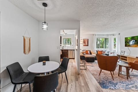 This space features a cozy dining nook with sleek black chairs and a modern pendant light. The living area offers a comfortable sofa, leather chairs, and a wall-mounted TV, with large windows filling the room with natural light, creating a fresh and invit