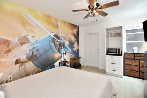 Sleeping area showing microwave oven, ceiling fan, dresser looking at a modern airplane theme painting on wall
