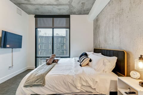Stylish bedroom with industrial accents, cozy bedding, flat-screen TV, and large windows offering natural light for a restful stay 🛏️🌆✨