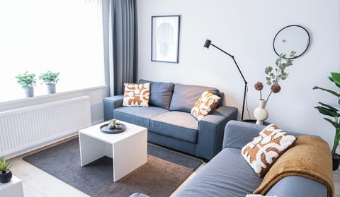 Stylish living room featuring two cozy gray sofas with warm-toned throws and patterned cushions. A sleek white coffee table, floor lamp, and decorative elements like potted plants and a modern wall mirror create an inviting atmosphere. Large windows allow natural light to enhance the bright and contemporary design.