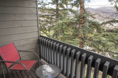 A small balcony with a red chair and round glass table, overlooking a scenic view of pine trees and distant hills.