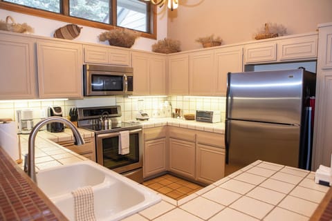 A modern kitchen with beige cabinets, stainless steel appliances, a double-basin sink, and a tiled countertop and backsplash. Decor items sit above the cabinets, and a window brings in natural light.