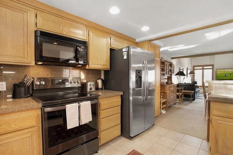 A modern kitchen featuring wooden cabinets, stainless steel appliances including a refrigerator and oven, a microwave above the oven, and a view into the adjacent living area.