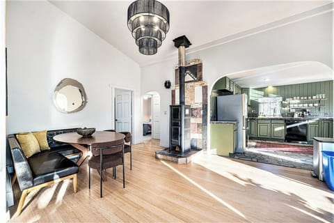 - Dine in a modern nook with tufted seating, sculptural chairs, and sunlit ambiance
- Unique tiled wood stove adds character and warmth to the open layout
- Olive green kitchen cabinets, floating shelves, and stone floors exude rustic elegance