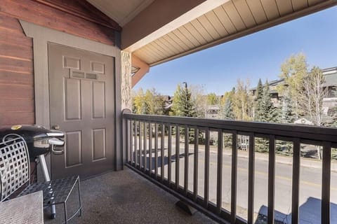 A small balcony with a grill, a chair, and a view of trees and a residential street. A closed door is on the left, and a railing runs along the edge of the balcony.