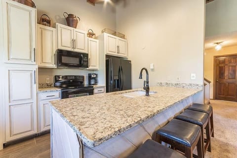 Modern kitchen with light-colored granite countertops, white cabinetry, black appliances, and black faucet on island counter with three dark brown stools.