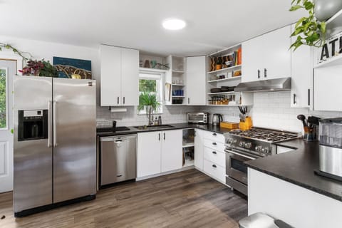 Modern kitchen with stainless steel appliances, white cabinets, a gas stove, double-door refrigerator, and wooden flooring. Potted plants and various kitchen items are visible on the countertops and shelves.