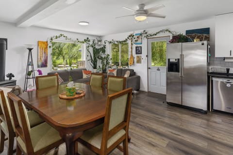 Dining and living area with wooden table and chairs, stainless steel refrigerator, couch, wall decorations, and various plants. Large windows and a glass door provide natural light.