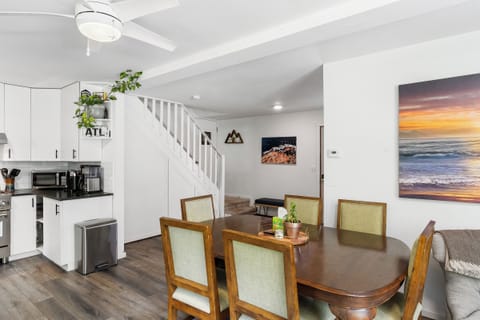 A dining area with a wooden table and six chairs, adjacent to a kitchen with white cabinets. A staircase leads to the upper floor, and ocean-themed artwork adorns the walls.