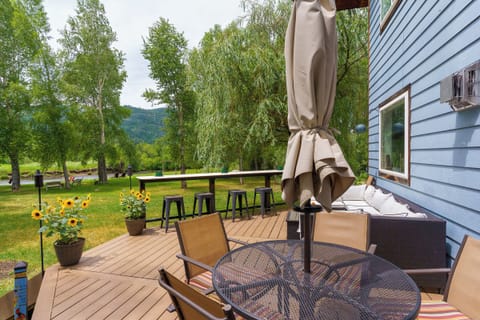 A backyard deck with a round table and chairs, a bar counter with stools, potted sunflowers, and an umbrella, adjacent to a blue house, with trees and grass in the background.