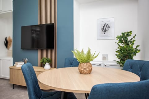 Stylish dining area with a modern wooden table, blue chairs, and a wall-mounted TV. Cozy, elegant, and inviting.