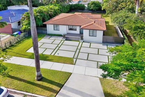 Aerial view reveals a unique property featuring geometric concrete pavers that create a captivating pattern leading to the home. The house features a classic barrel tile roof and is surrounded by lush tropical landscaping.