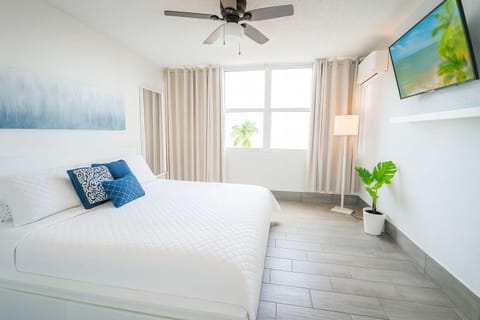 Bright and elegant king bedroom with ocean-inspired decor, crisp white bedding, and plenty of natural light. Enjoy a relaxing atmosphere with a wall-mounted TV and modern tropical touches for a serene island escape.