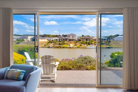 A cozy indoor space with a large sliding door that offers a scenic view of a waterfront area with houses in the background. Inside, there is a comfortable blue armchair with a pillow, and outside on the patio, there is a white Adirondack chair, surrounded