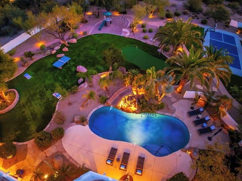 An aerial view captures the property's impressive resort-style amenities at twilight, featuring a sparkling pool, putting green, tennis court, and lush landscaped grounds illuminated by ambient lighting.