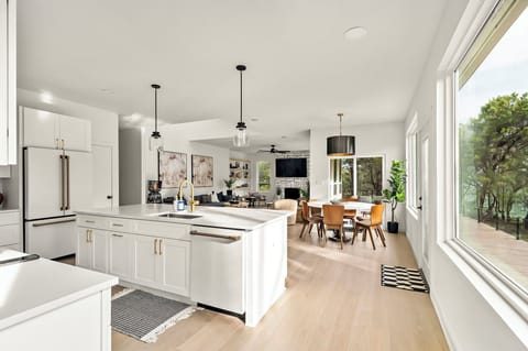 Bright open-concept kitchen and dining area featuring a large white island with gold fixtures, pendant lighting, modern white cabinetry, a farmhouse sink, wooden dining table with chairs, and a cozy living space with a fireplace and mounted TV.