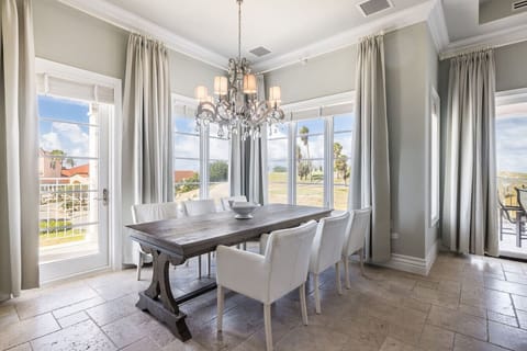 A bright dining area with a rustic wooden table, white chairs, a stylish chandelier, and large windows offering scenic views.